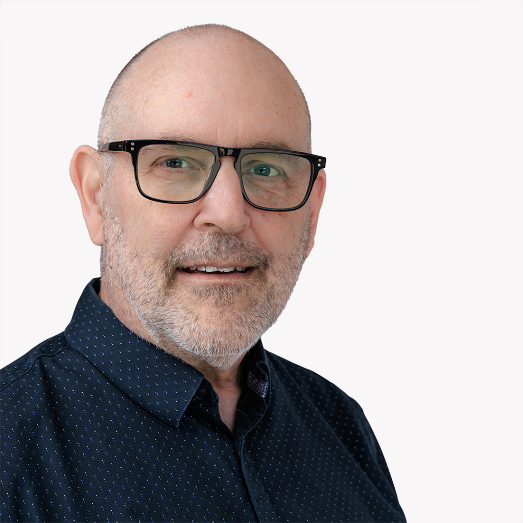 Close up corporate headshot of a man with glasses and a neatly trimmed beard wearing a dark button down shirt against a clean light background, presenting a professional and approachable appearance.