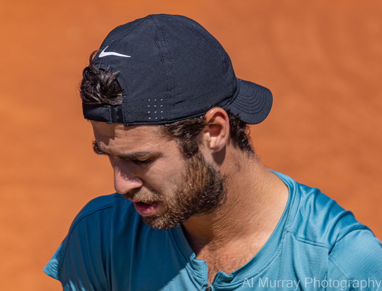 Close up of a tennis player in a blue shirt and backward cap looking down between points on a clay court, highlighting a focused and candid moment often seen in tennis images.