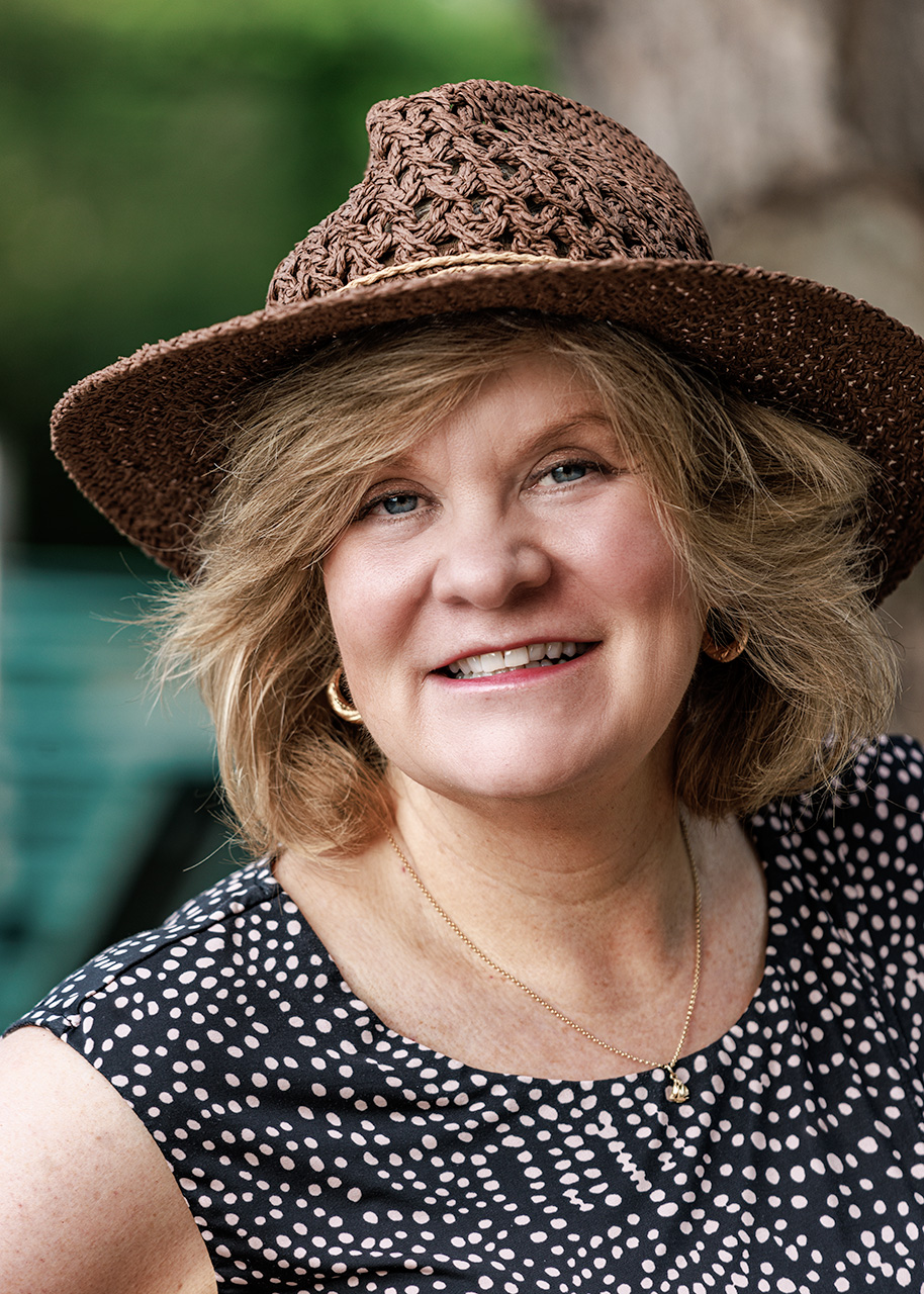 Smiling woman wearing a woven brown hat and patterned sleeveless top poses outdoors near water, conveying a warm and approachable look ideal for personal branding.