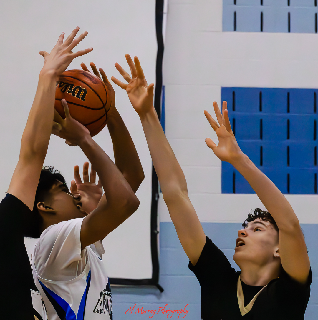 Two players jump with arms raised to contest a basketball near the hoop during a local basketball game in an indoor gym, capturing a tense defensive moment. Blue wall padding and a blurred court setting frame the action.